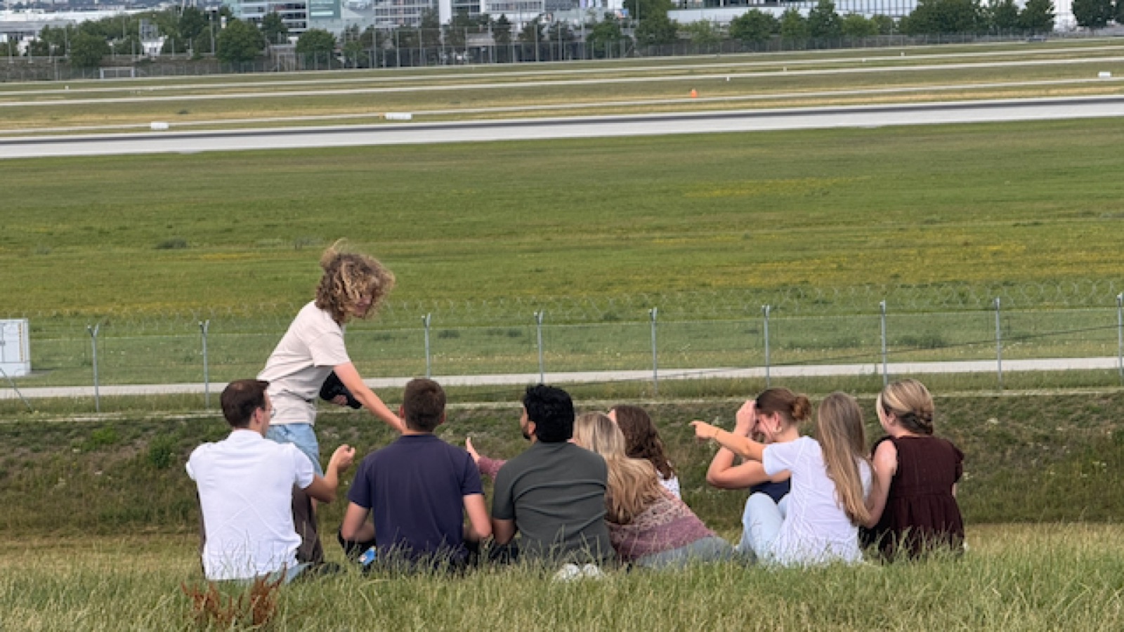 Friends on the grass by the Munich Airport runway fence, Luca climbing for a better view